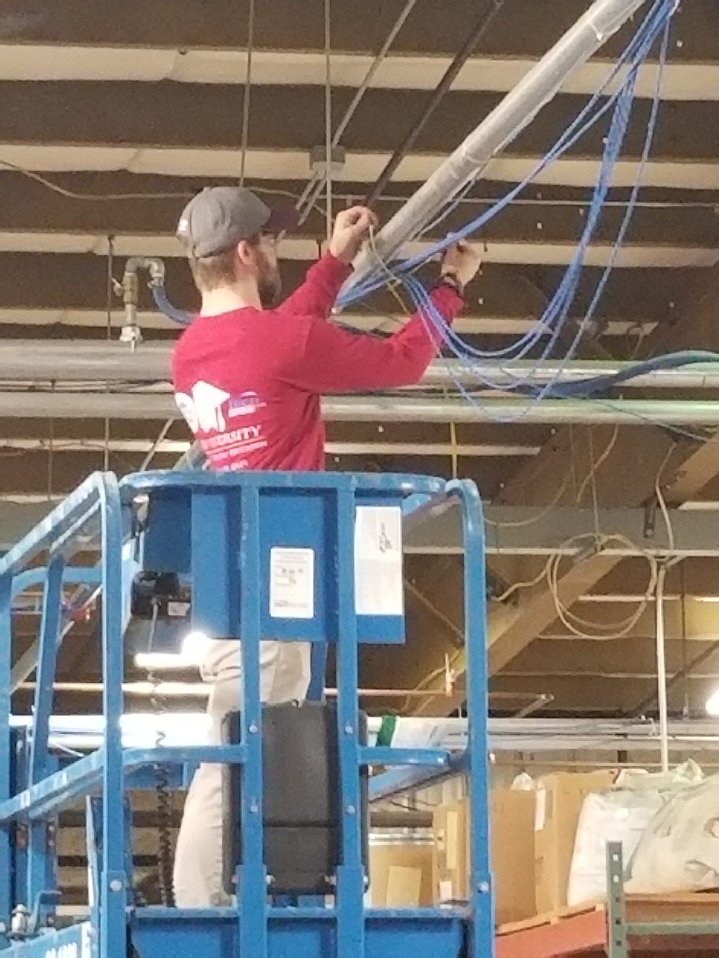 A technician on a scissor lift pulling bundles of blue network cable into overhead cable tray, in a building with the ceiling still open