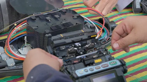 A technician aligning fiber strands in a fusion splicer in the field, with colour-coded fiber and a splice tray alongside