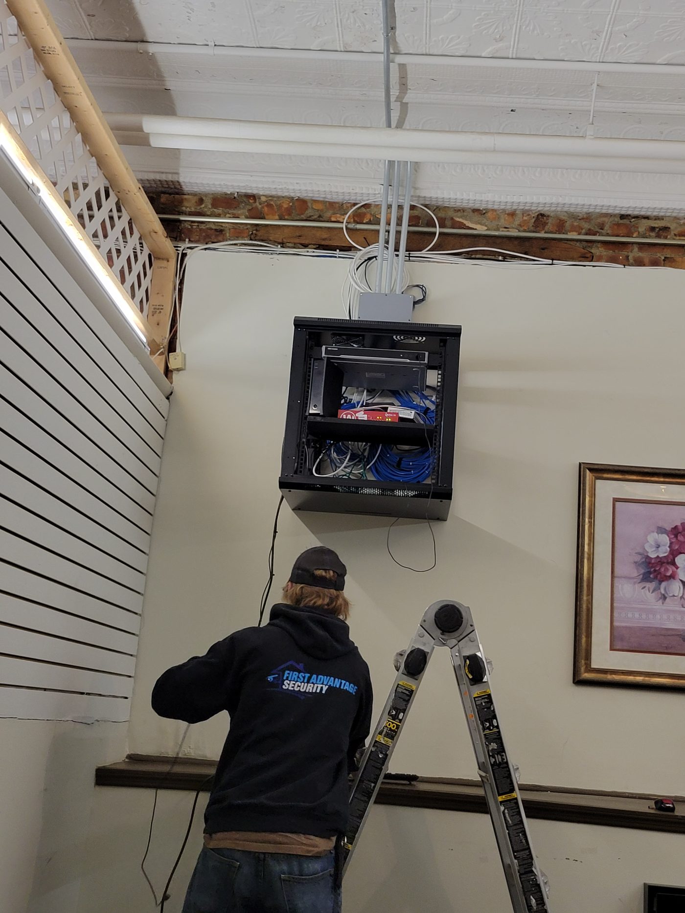 A First Advantage technician on a step ladder beside a wall-mounted network rack holding a network switch and an NVR, with dressed blue patch cables, in a building with a pressed tin ceiling and exposed brick