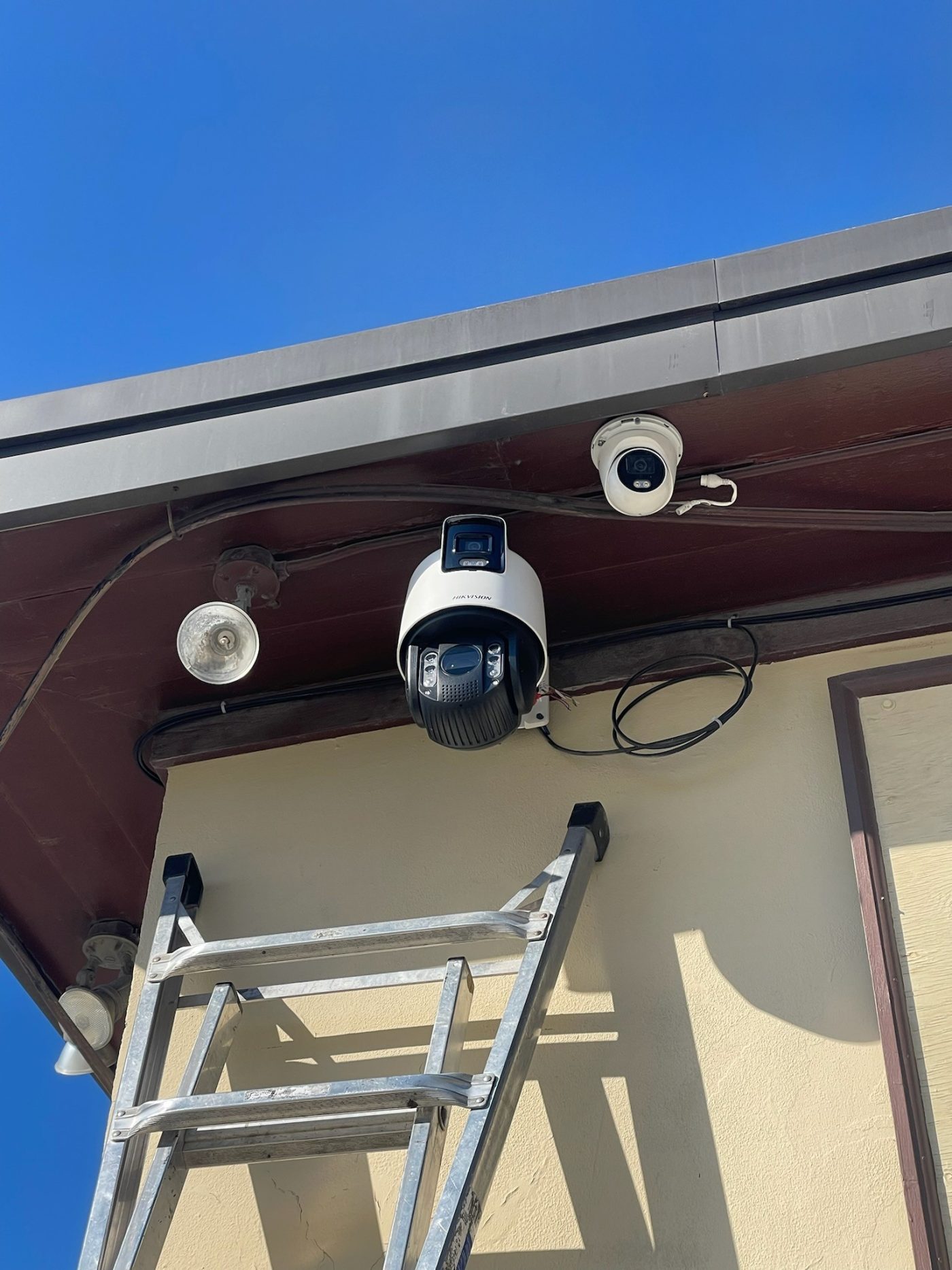 A pan-tilt-zoom camera and a fixed turret camera mounted under the soffit of a building, with cabling run along the eave and a service loop coiled beside them