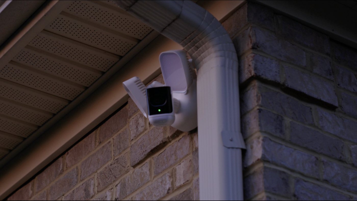 A floodlight security camera mounted under the soffit of a brick house at dusk, its status light showing green